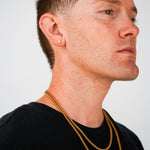 A close-up side profile of a man with short brown hair, wearing two gold chain necklaces and a small gold stud earring, against a plain light gray background.