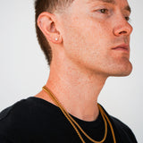 A close-up side profile of a man with short brown hair, wearing two gold chain necklaces and a small gold stud earring, against a plain light gray background.