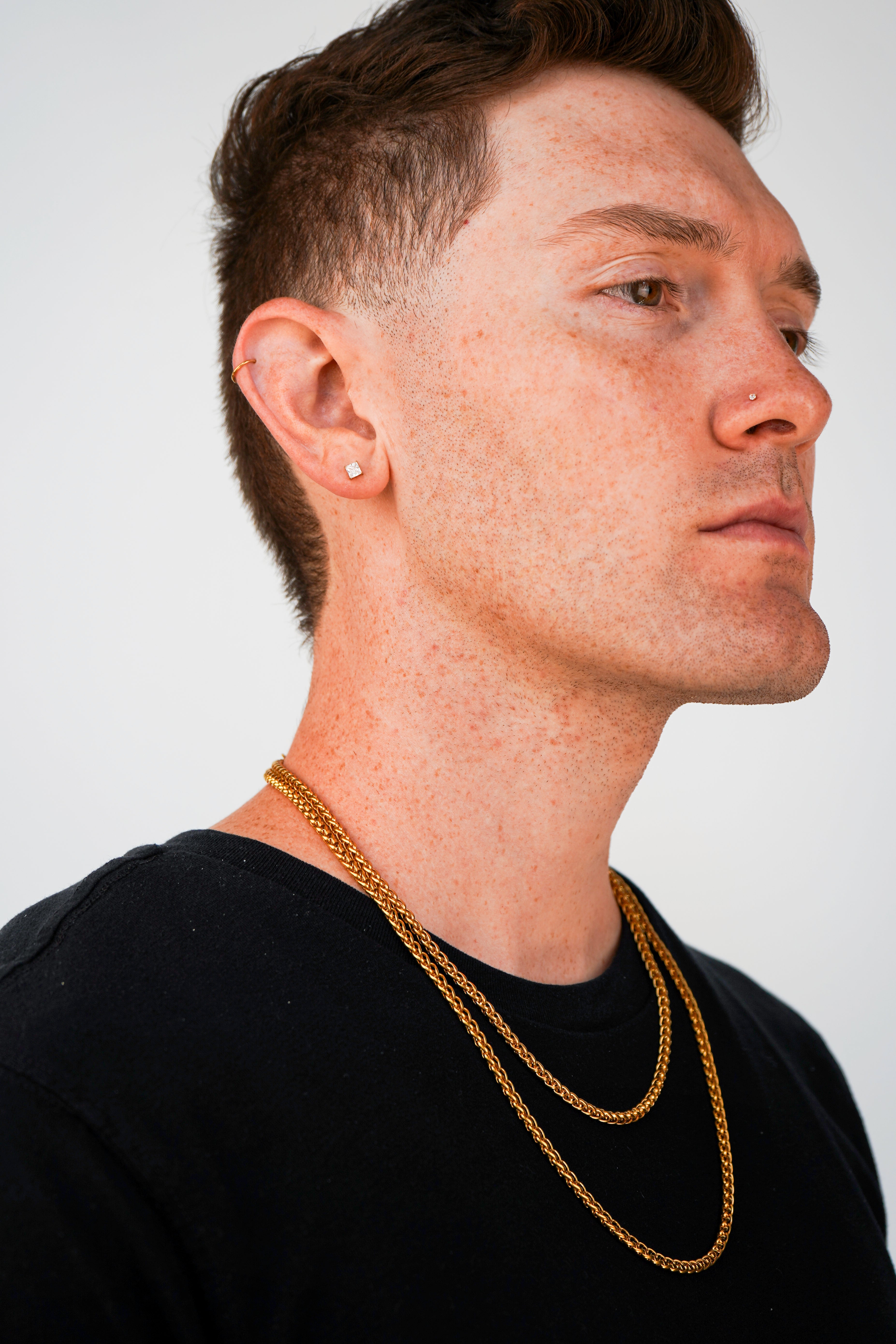 A close-up side profile of a man with short brown hair, wearing two gold chain necklaces and a small gold stud earring, against a plain light gray background.