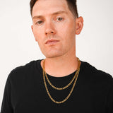 A man with short brown hair and tattoos wearing a black t-shirt and two layered gold chain necklaces, photographed against a plain light gray background.