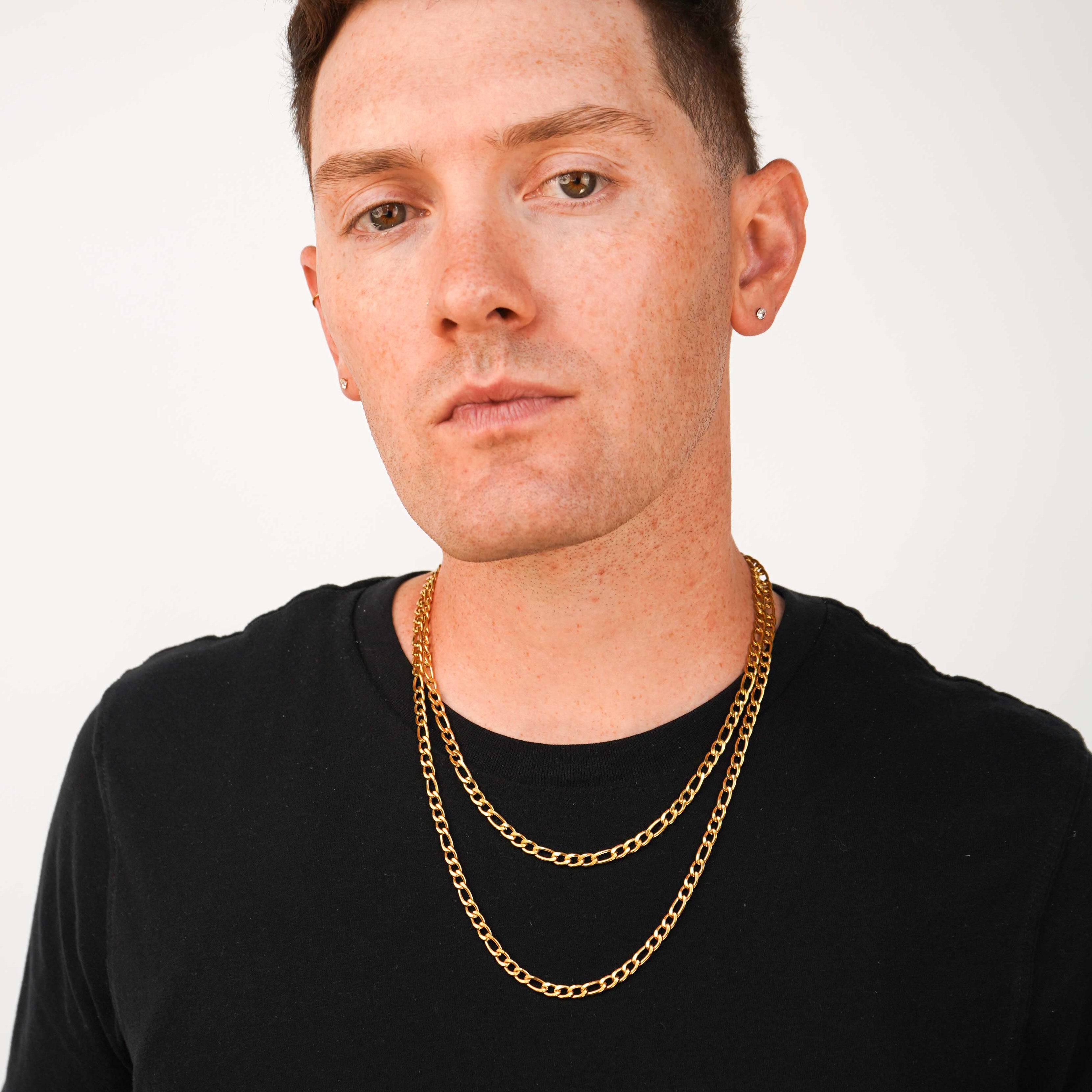 A man with short brown hair and tattoos wearing a black t-shirt and two layered gold chain necklaces, photographed against a plain light gray background.