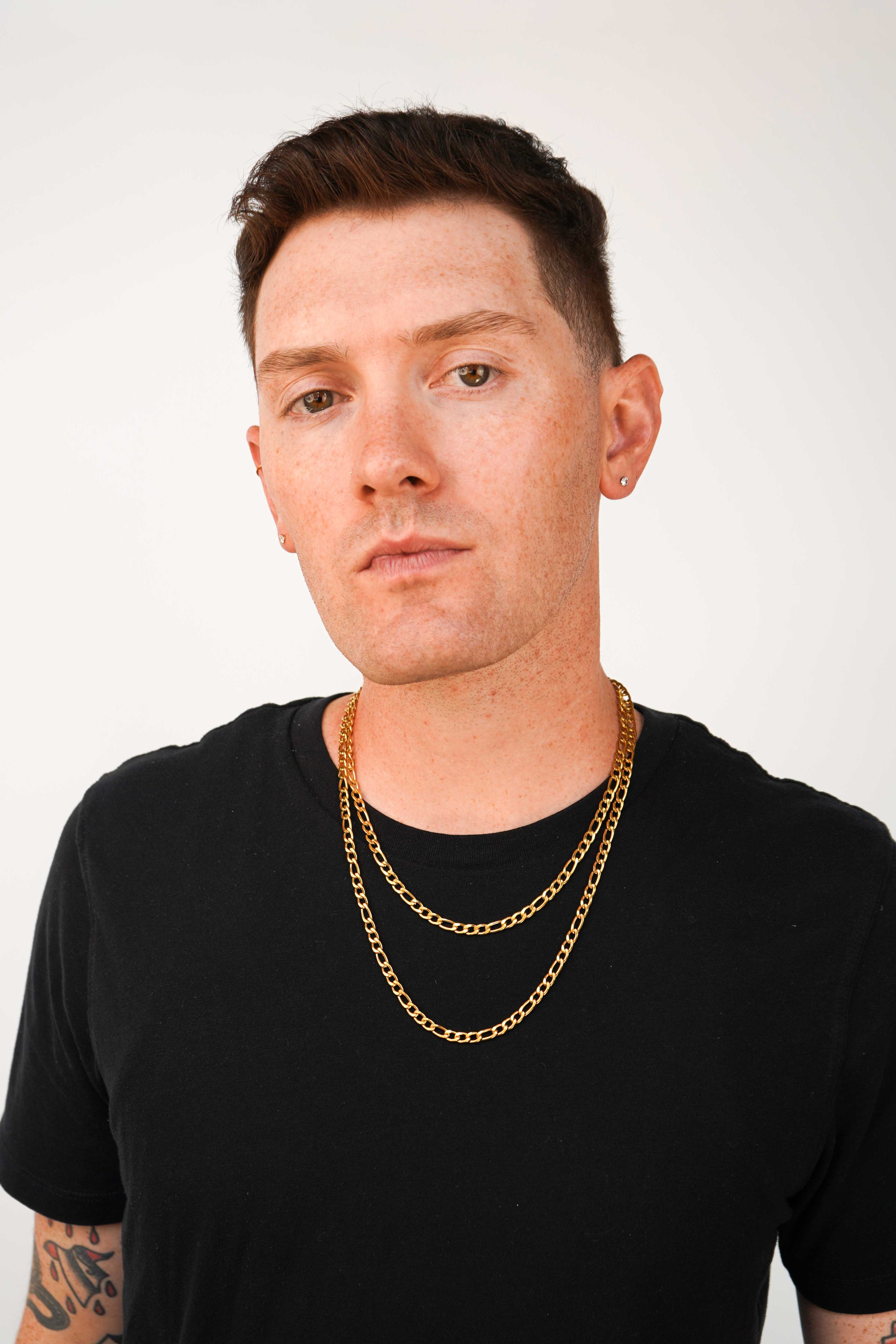 A man with short brown hair and tattoos wearing a black t-shirt and two layered gold chain necklaces, photographed against a plain light gray background.