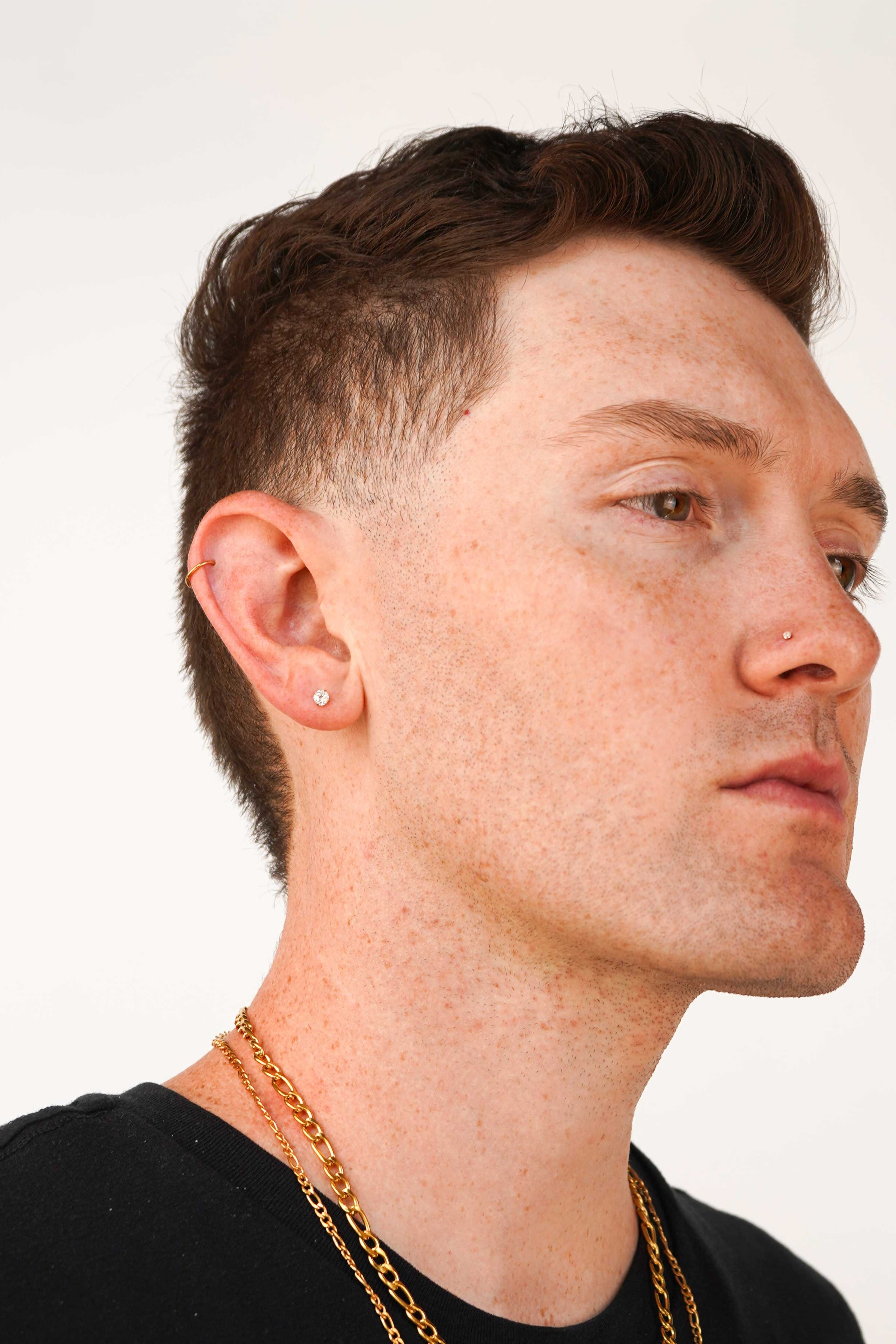 Close-up of a man's ear wearing a small gold and clear CZ stud earring, with additional ear piercings and a gold chain necklace visible against a plain light background.