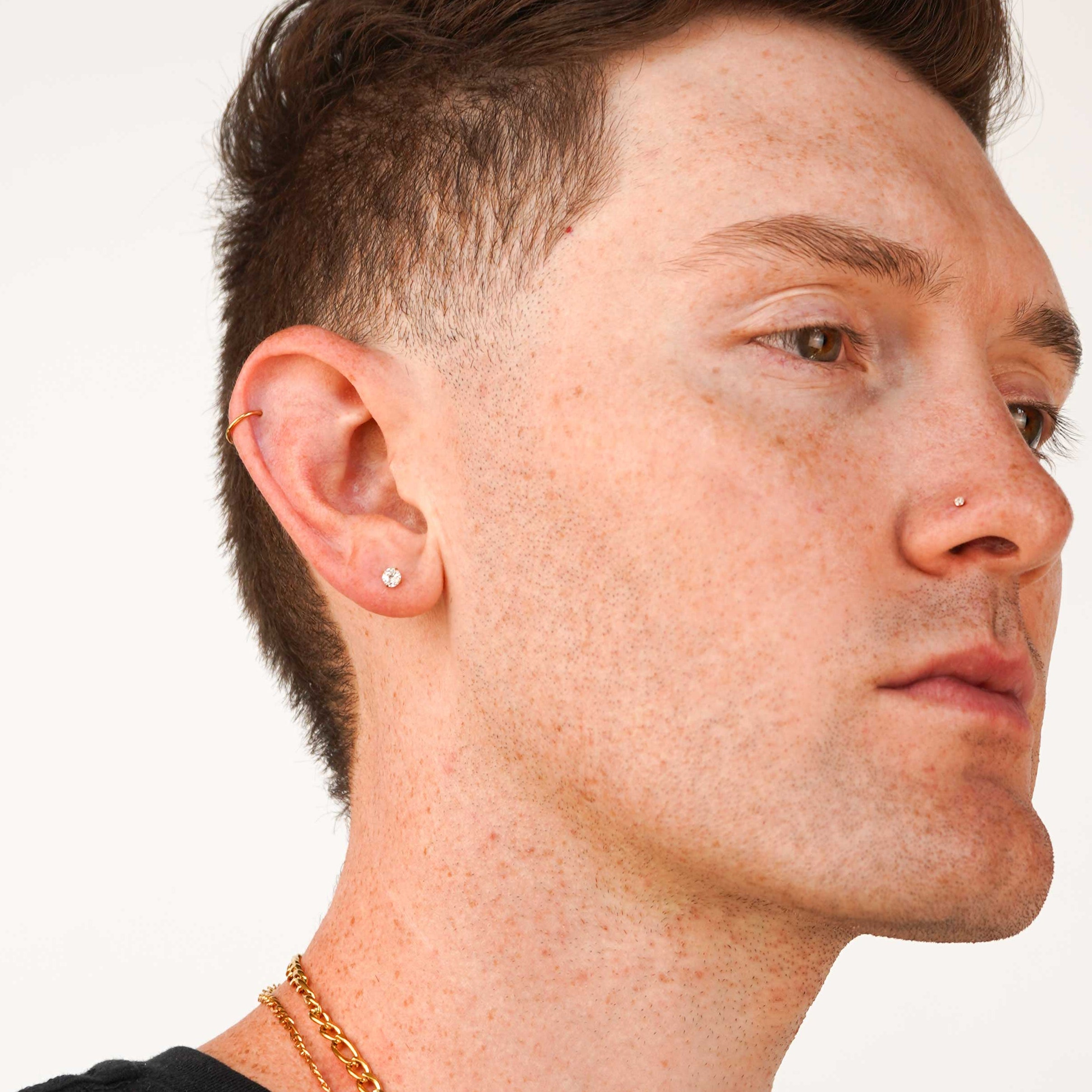 Close-up of a man's ear wearing a small gold and clear CZ stud earring, with additional ear piercings and a gold chain necklace visible against a plain light background.