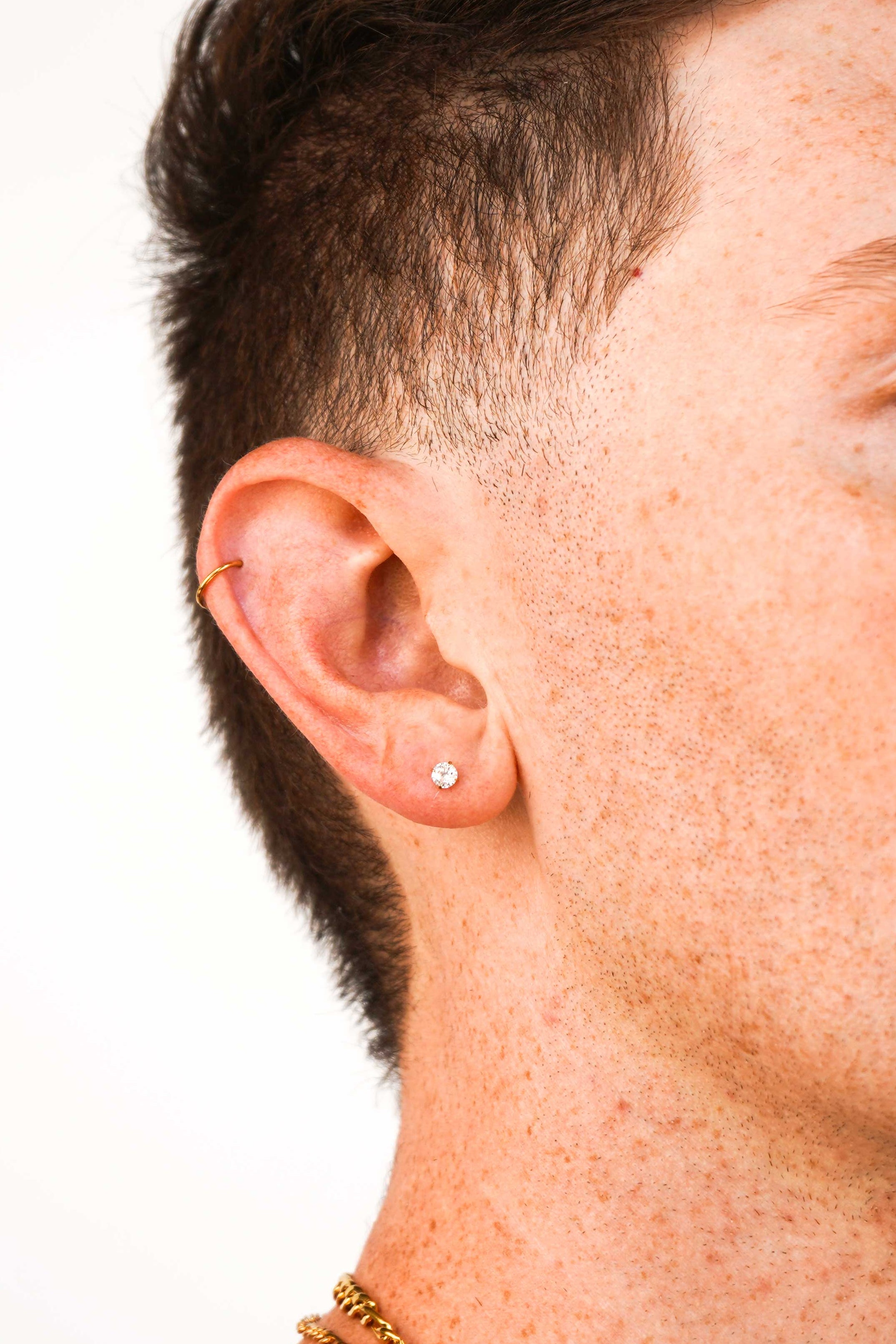 A close-up side profile of a man with short dark hair, wearing a small round cz stud and small helix hoop earring in his left ear and a gold chain necklace, against a plain light gray background.