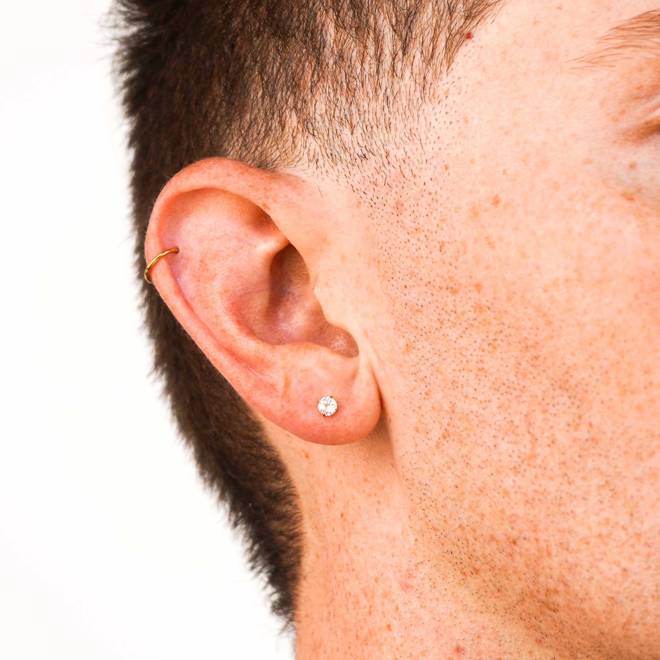 A close-up side profile of a man with short dark hair, wearing a small round cz stud and small helix hoop earring in his left ear and a gold chain necklace, against a plain light gray background.
