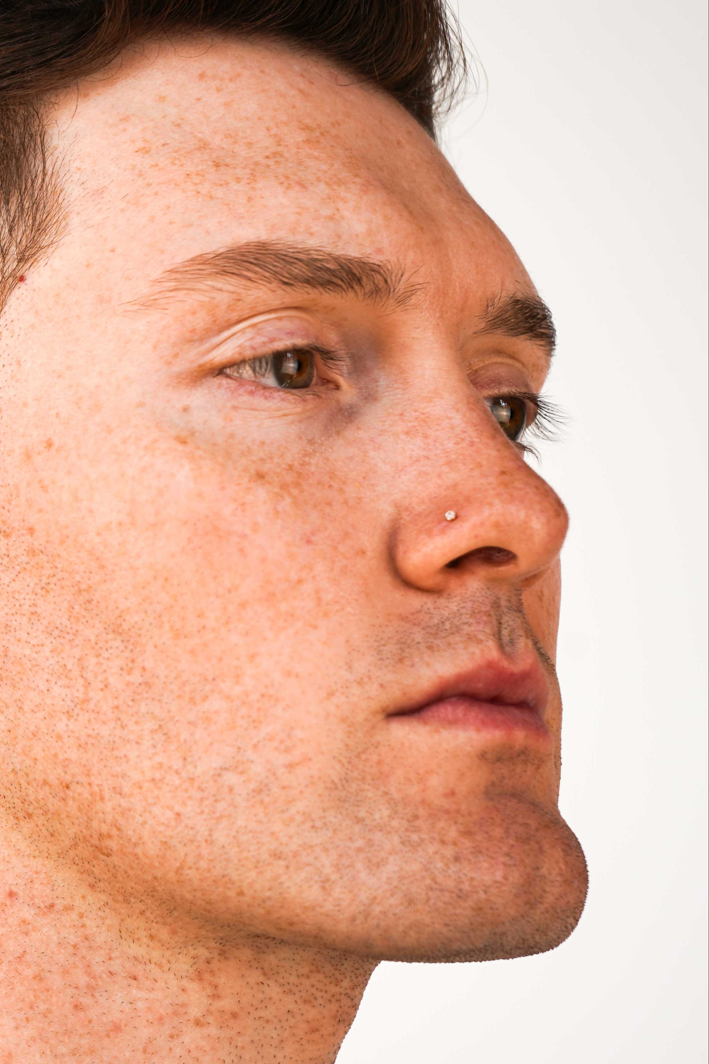 A close-up side profile of a man with short brown hair, wearing a clear round CZ stud earring in his nose, against a plain white background.
