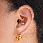 A close-up of a person's ear wearing a gold huggie hoop earring and a matching small drop earring with a dark round stone, plus a daith hoop, against a light neutral background.