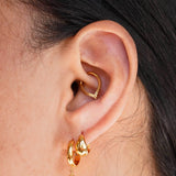 A close-up of a person's ear wearing a gold huggie hoop earring and a matching small drop earring with a dark round stone, plus a daith hoop, against a light neutral background.