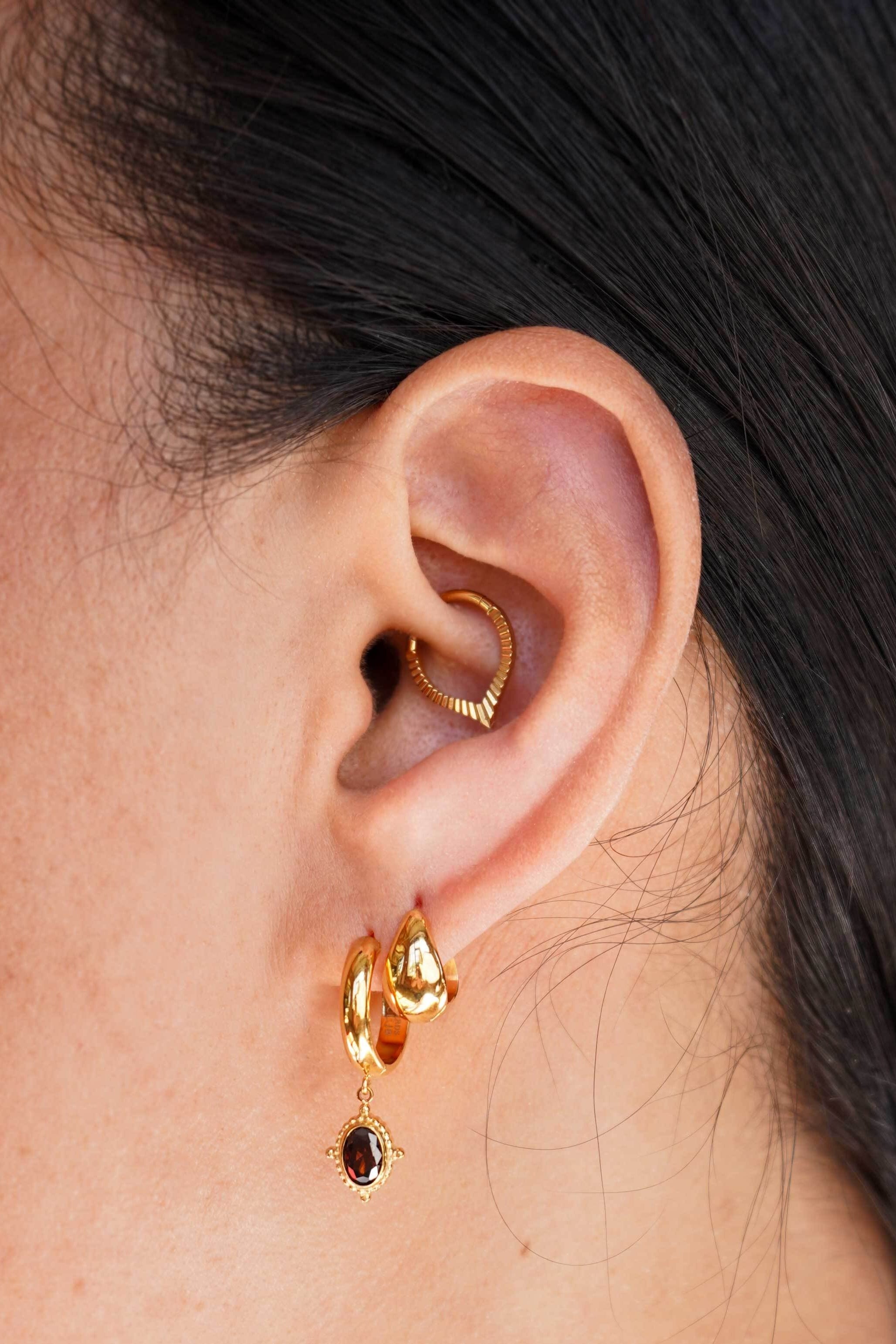 A close-up of a person's ear wearing a gold huggie hoop earring and a matching small drop earring with a dark round stone, plus a daith hoop, against a light neutral background.