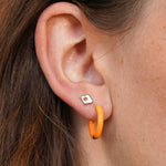 Close-up of an ear with a silver, white, and orange enamel flat back stud earring and orange hoop earring 