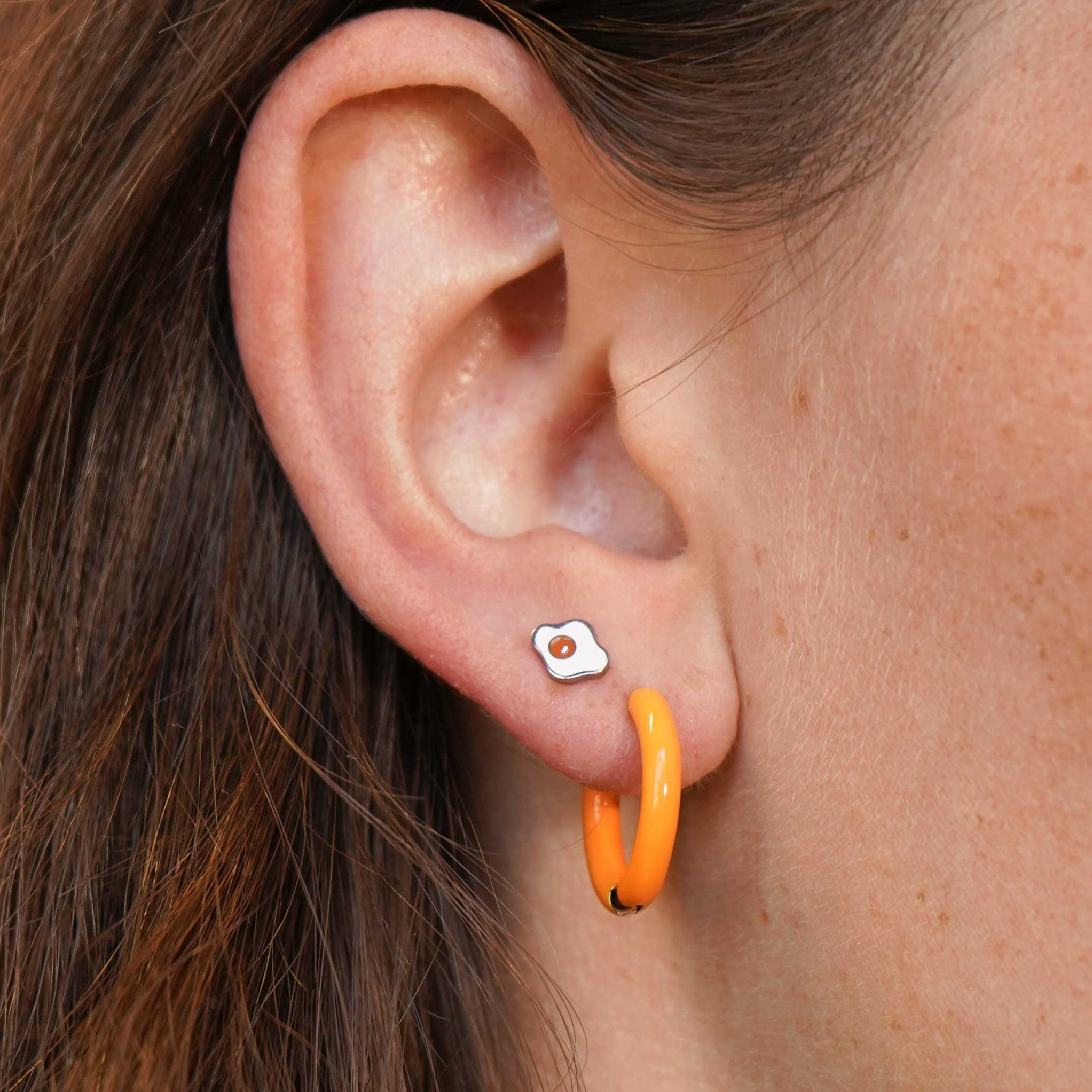 Close-up of an ear with a silver, white, and orange enamel flat back stud earring and orange hoop earring 
