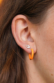 Close-up of an ear with a silver, white, and orange enamel flat back stud earring and orange hoop earring 
