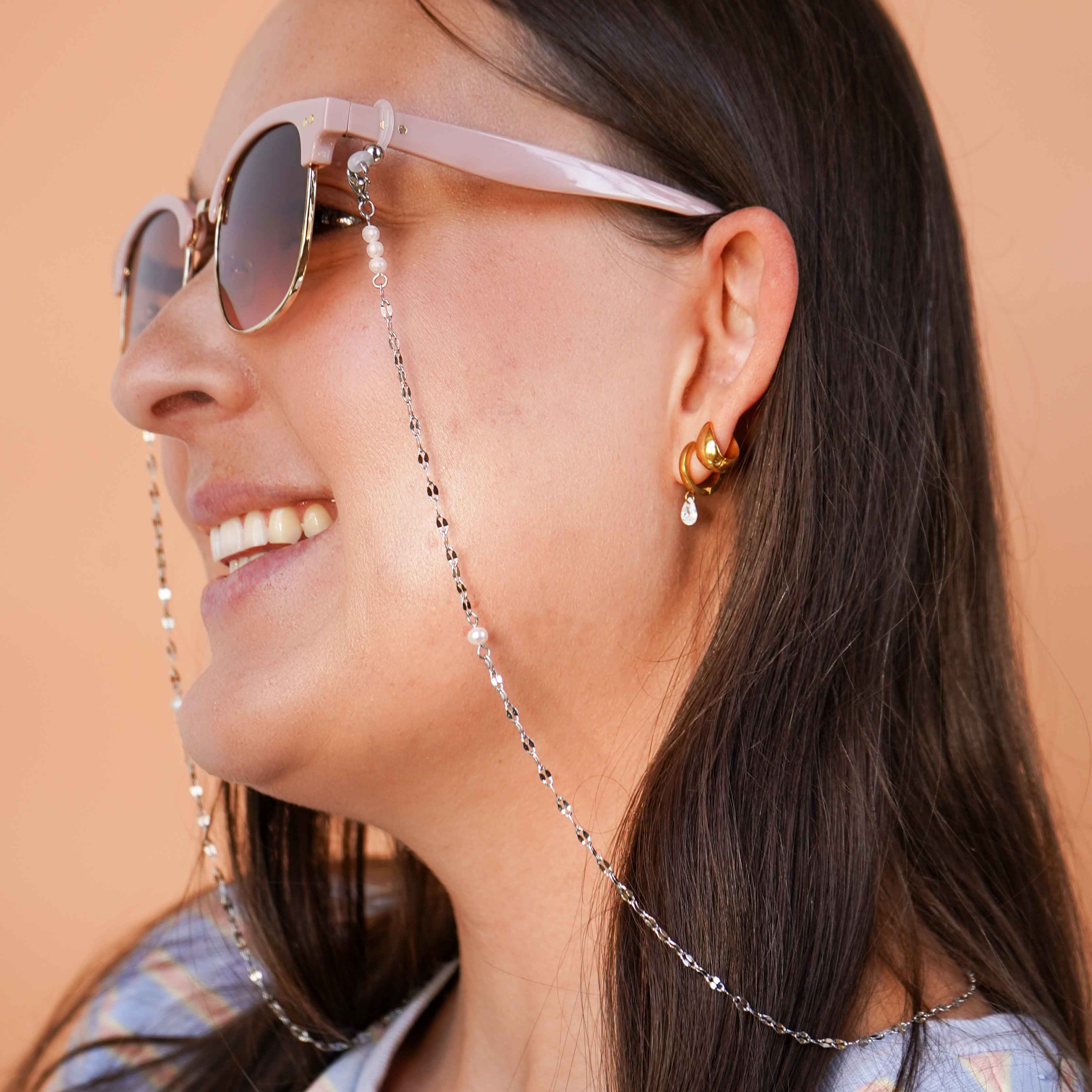 Woman wearing sunglasses with a silver chain, smiling against a beige background.