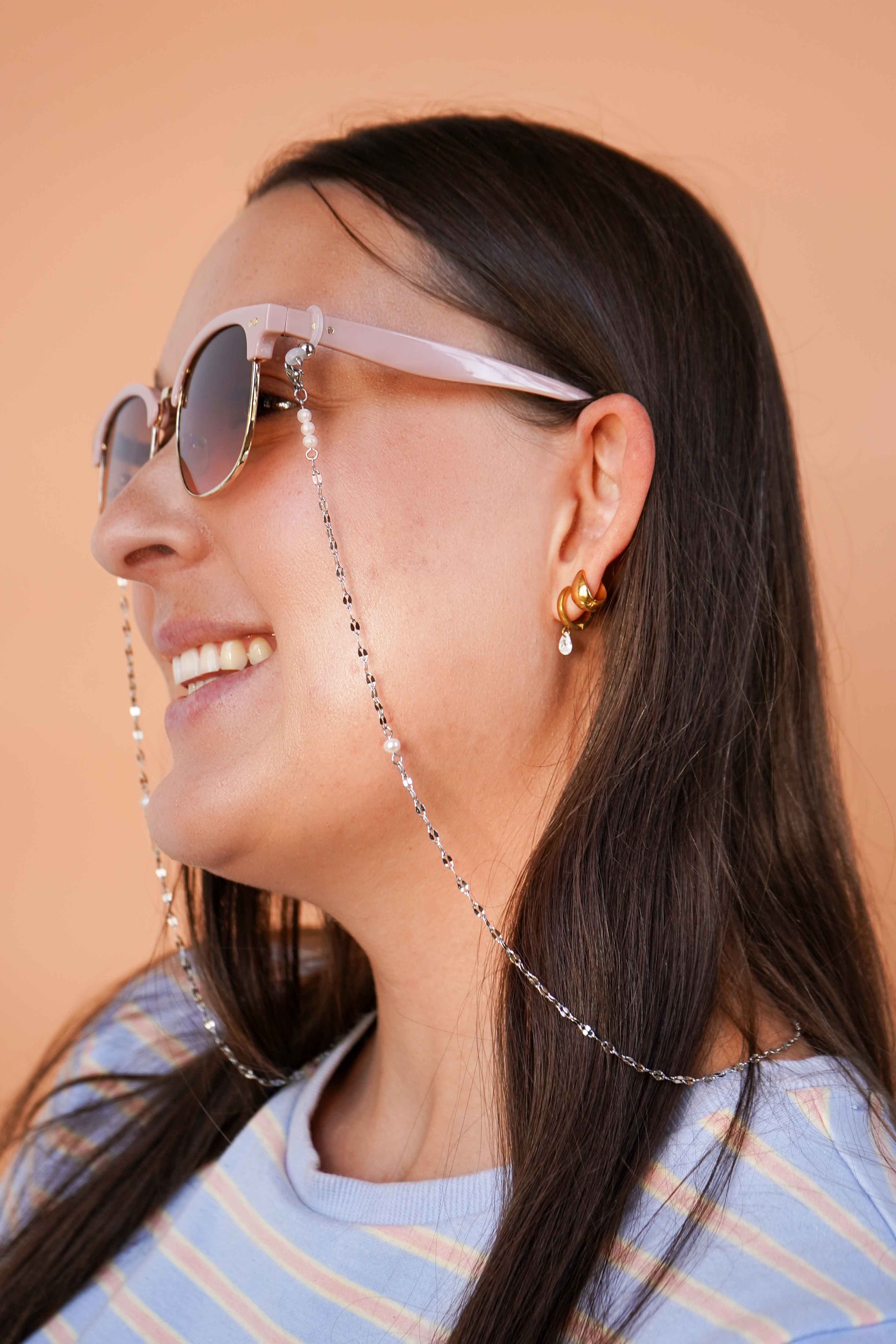 Woman wearing sunglasses with a silver chain, smiling against a beige background.