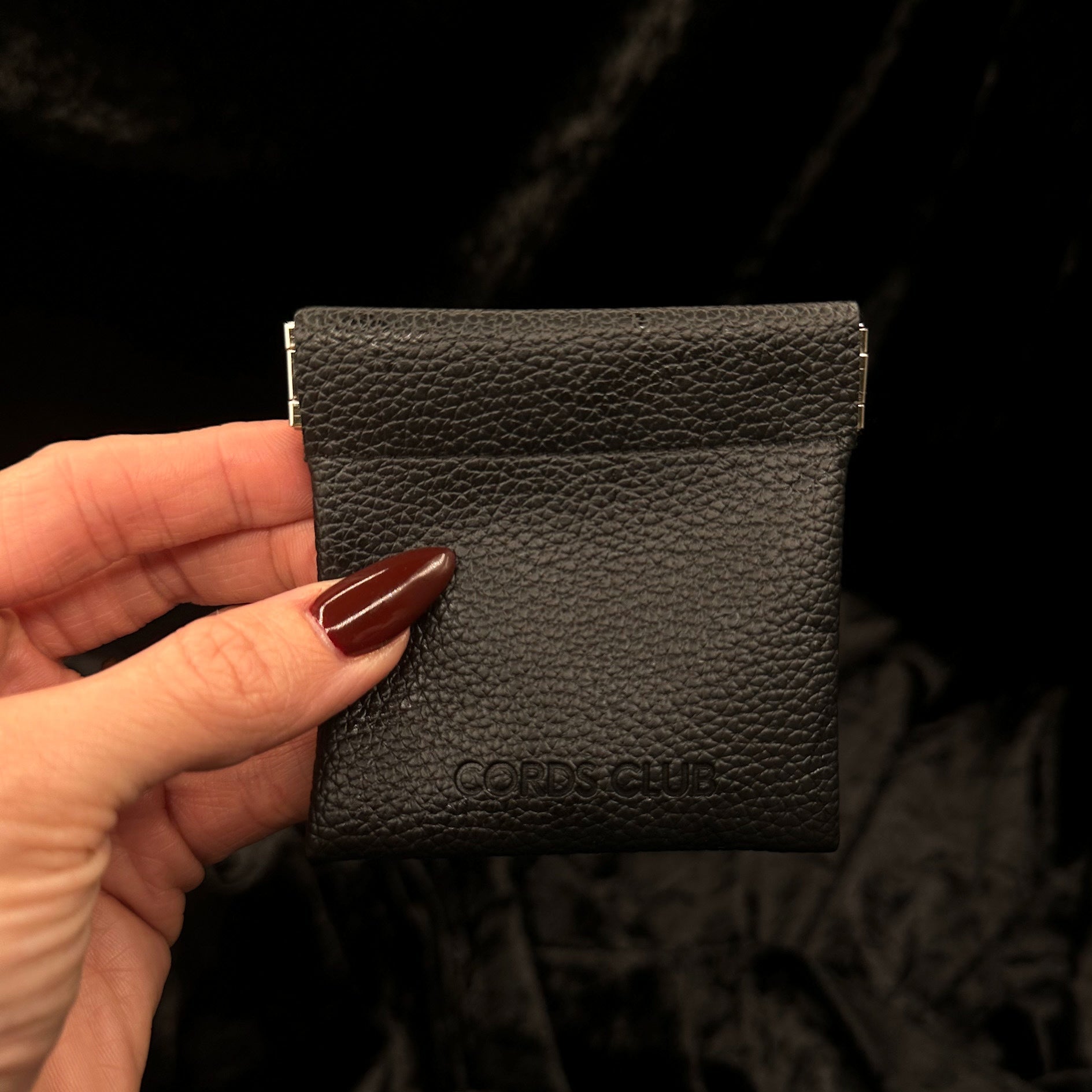 Hand holding a black faux leather pouch with 'Cords Club' branding against a dark background