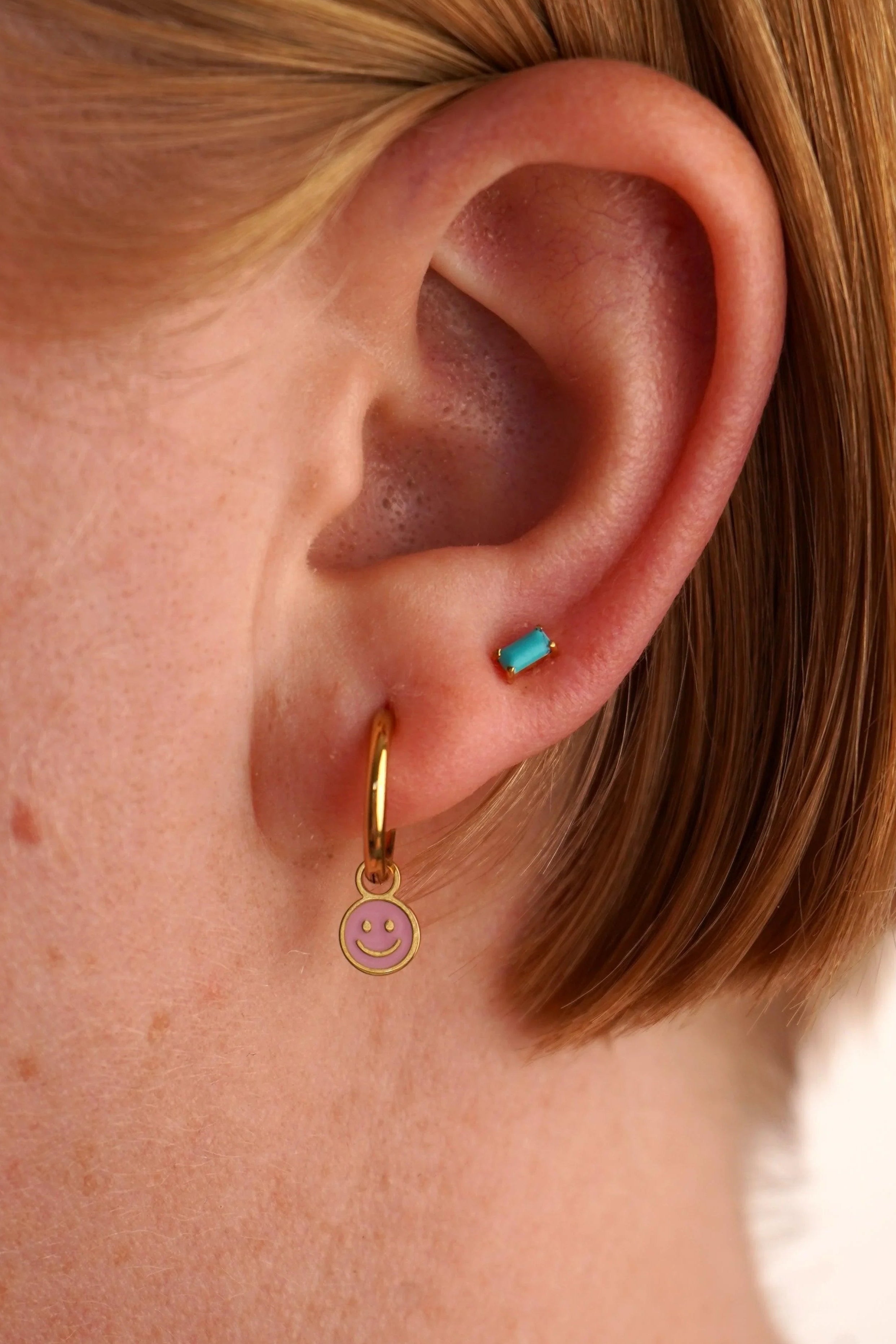 Close-up of an ear with a gold hoop earring featuring a smiley face charm.