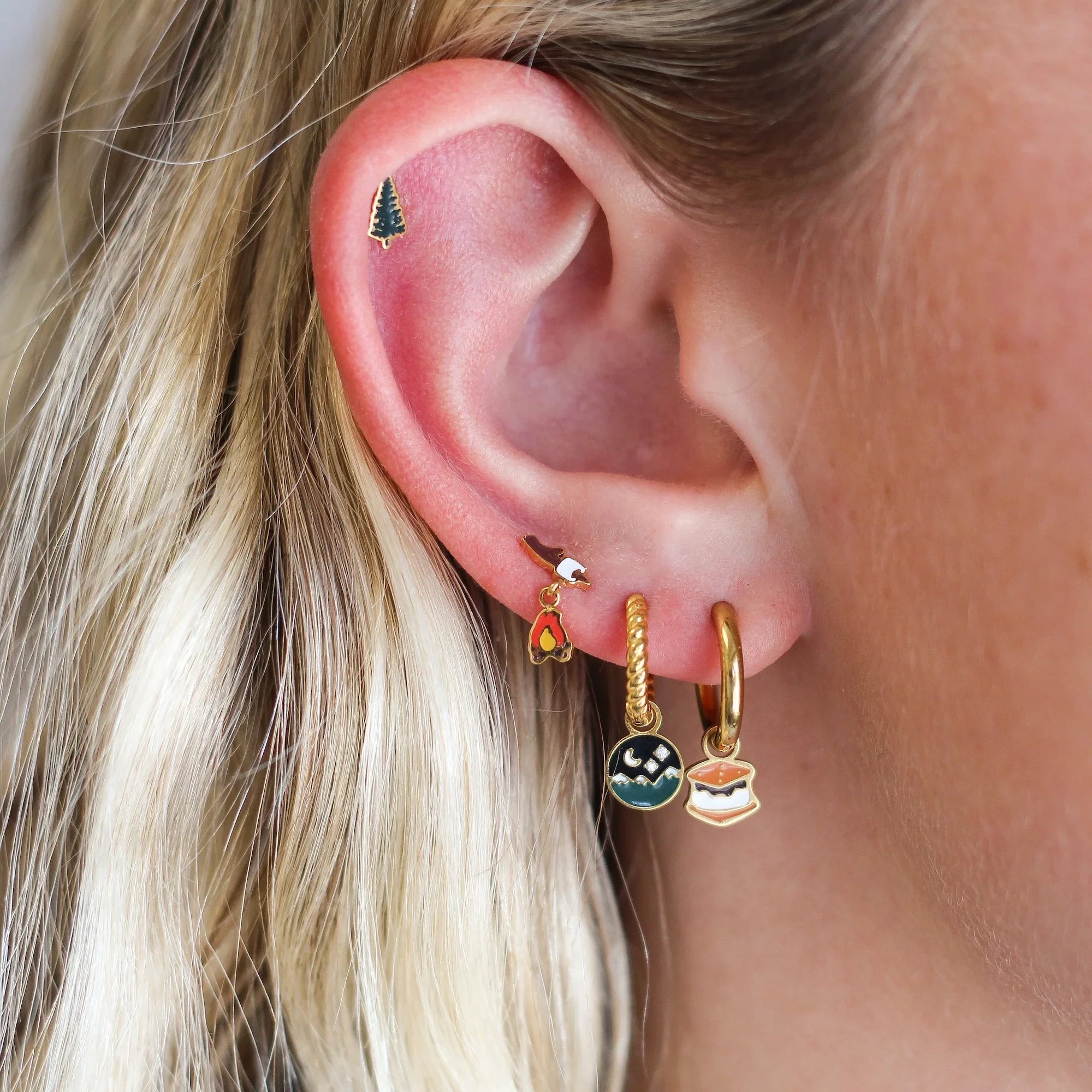Close up of an ear with multiple piercings, featuring a green pine tree stud and a marshmallow on a stick over a dangled campfire stud, plus 2 gold hoops, 1 with a s'mores charm and the other with  round night sky scene charm.