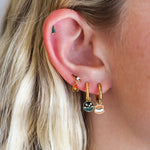 Close up of an ear with multiple piercings, featuring a green pine tree stud and a marshmallow on a stick over a dangled campfire stud, plus 2 gold hoops, 1 with a s'mores charm and the other with  round night sky scene charm.