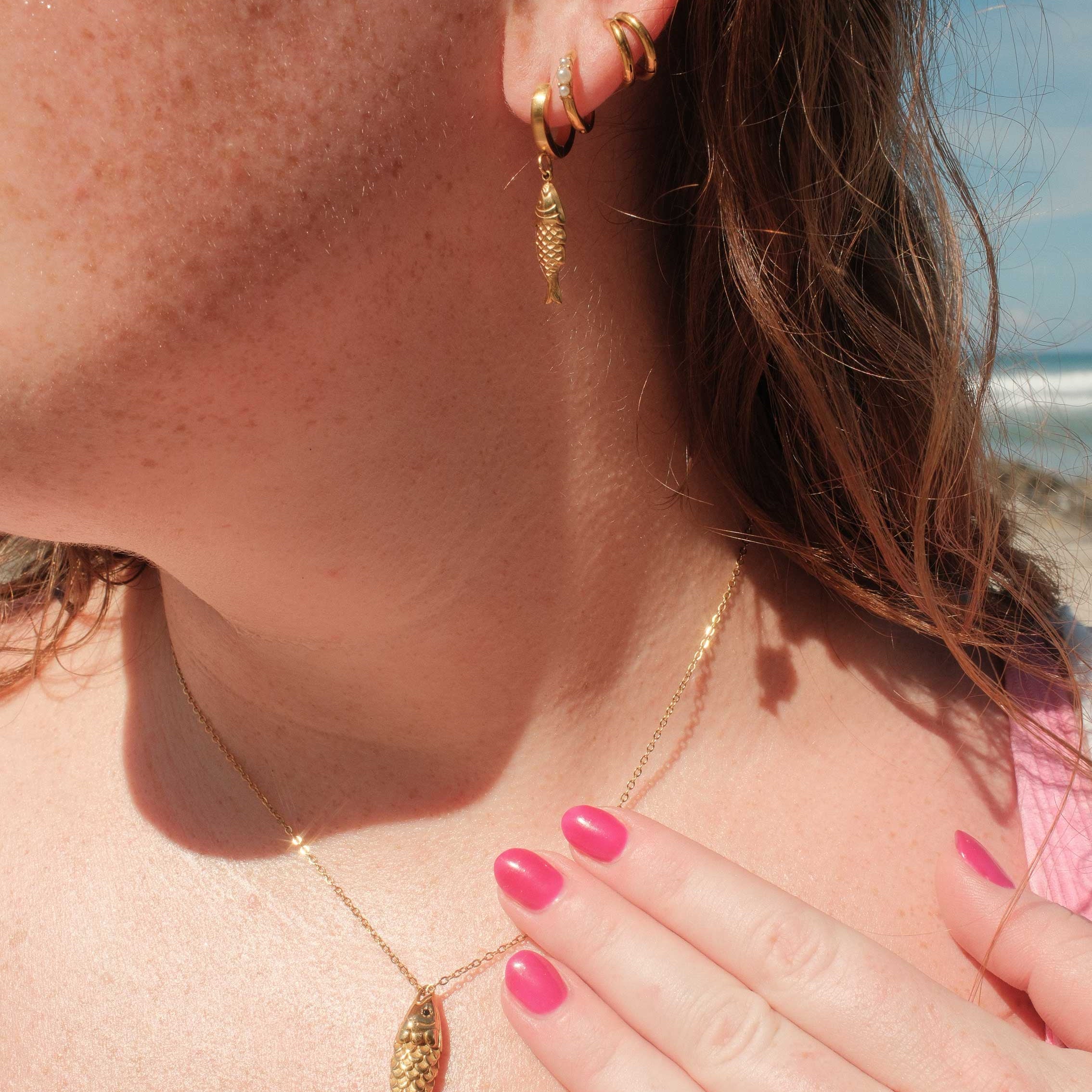 Close-up of a person wearing gold hoop earrings and a gold necklace with a beach in the background