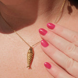 Gold necklace with a fish pendant worn by a person with pink nail polish.