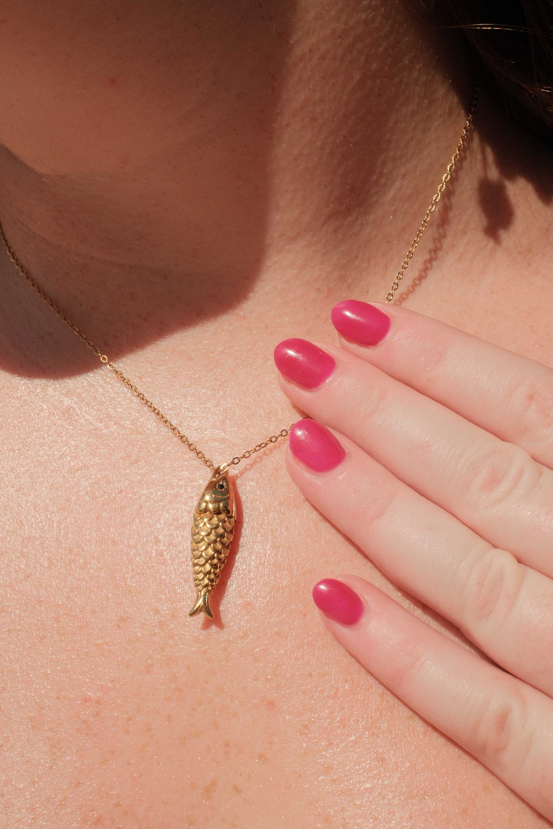 Gold necklace with a fish pendant worn by a person with pink nail polish.