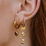 A close-up of a person's ear wearing a set of gold hoop earrings with dangling geometric charms, including a small dark oval stone and stacked cube-like drops.