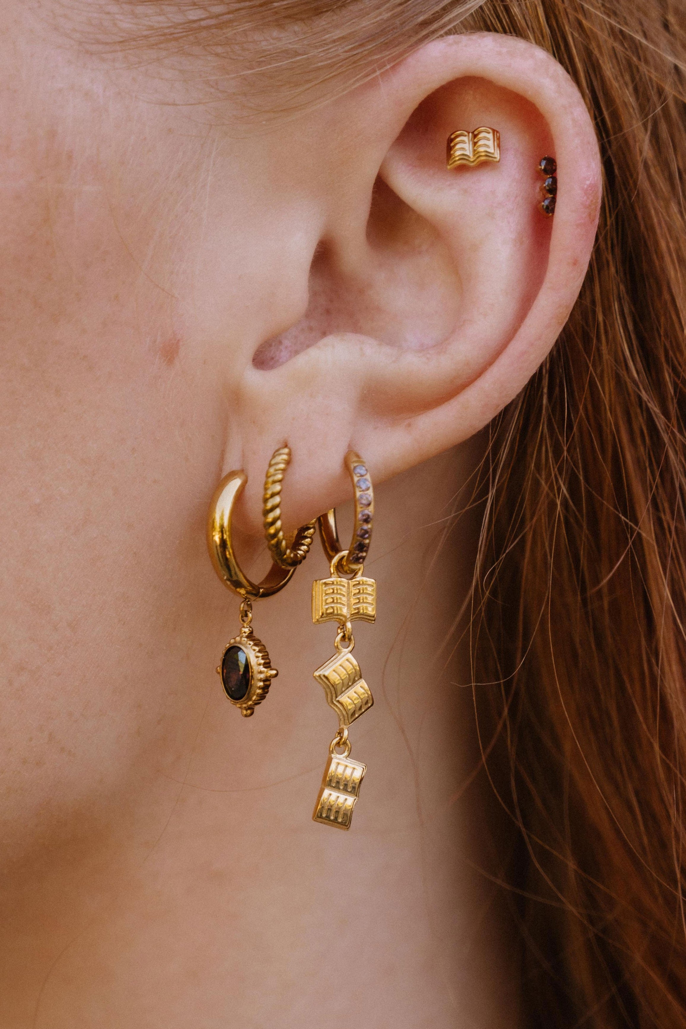 A close-up of a person's ear wearing a set of gold hoop earrings with dangling geometric charms, including a small dark oval stone and stacked cube-like drops.