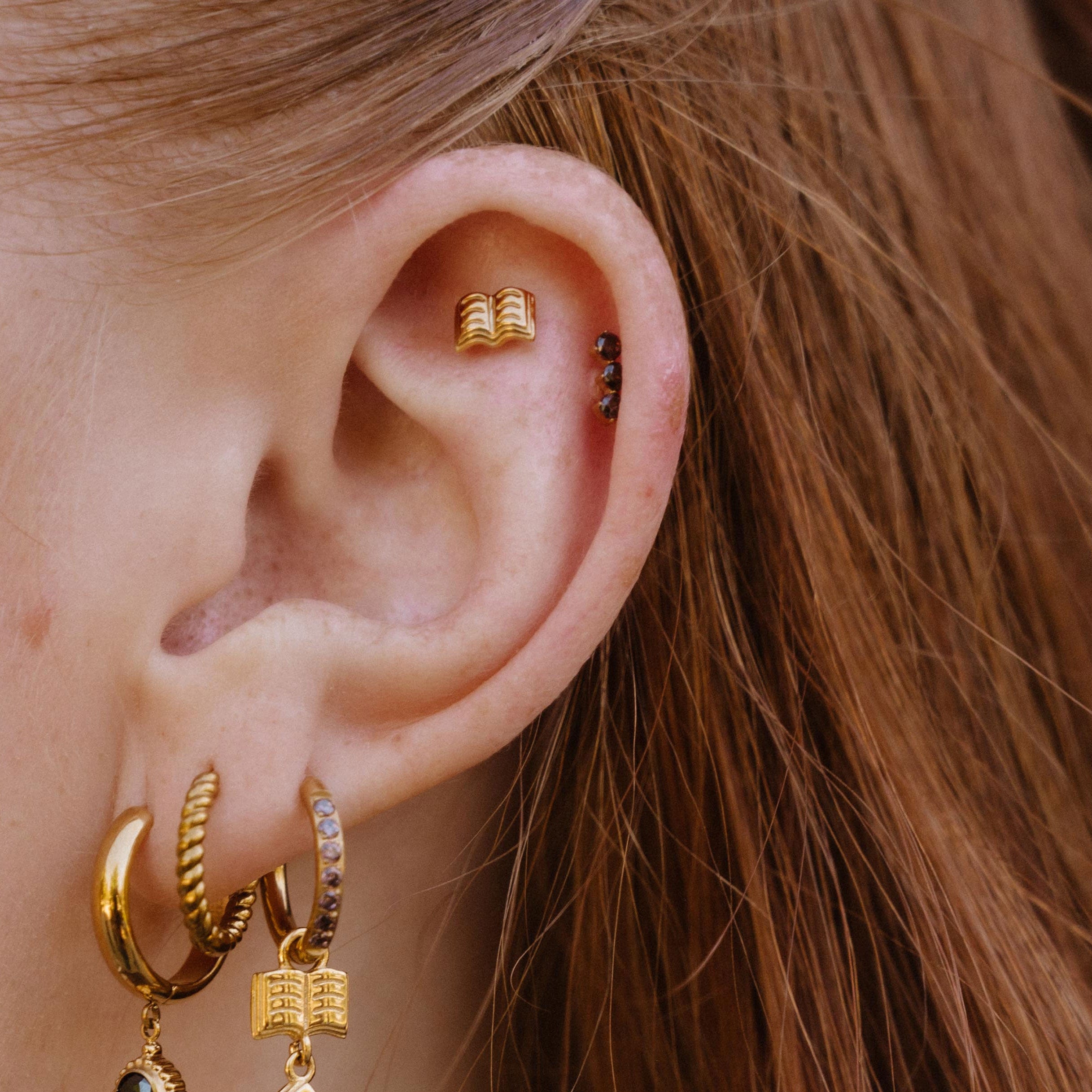 Close-up of a person's ear showcasing multiple gold piercings, including a small gold book charm earring with a dark stone in the lobe, a gold hoop, and a gold cartouche hoop with a small gold book charm.