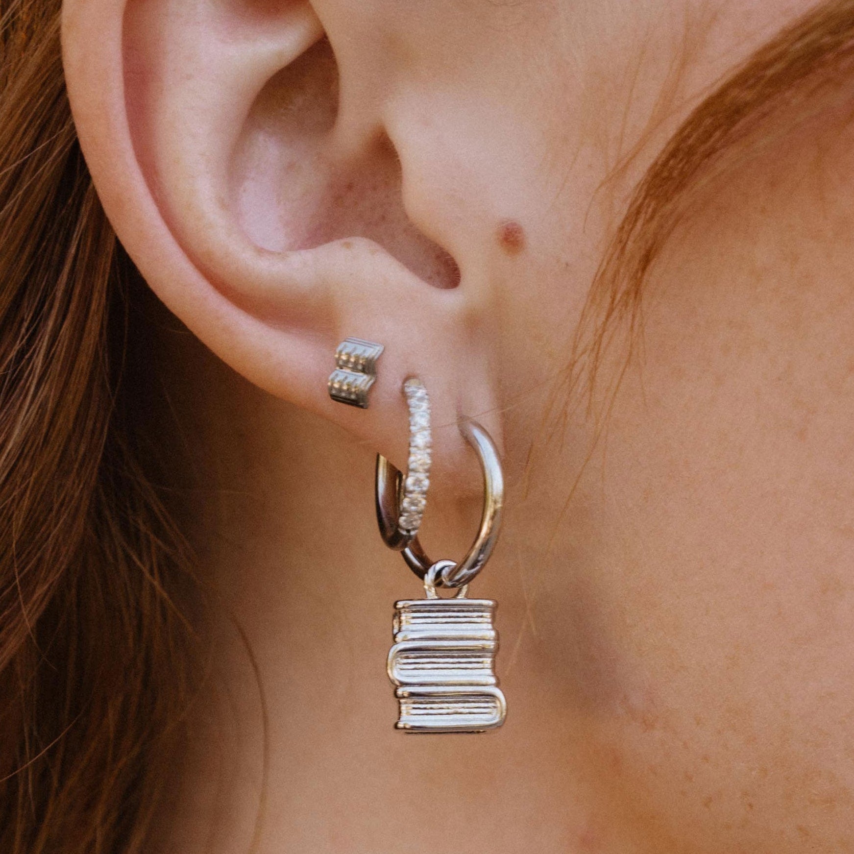 A close-up of a person's ear wearing multiple silver earrings, including a small stud, a huggie hoop with tiny clear stones, and a larger hoop with a dangling stacked books charm.