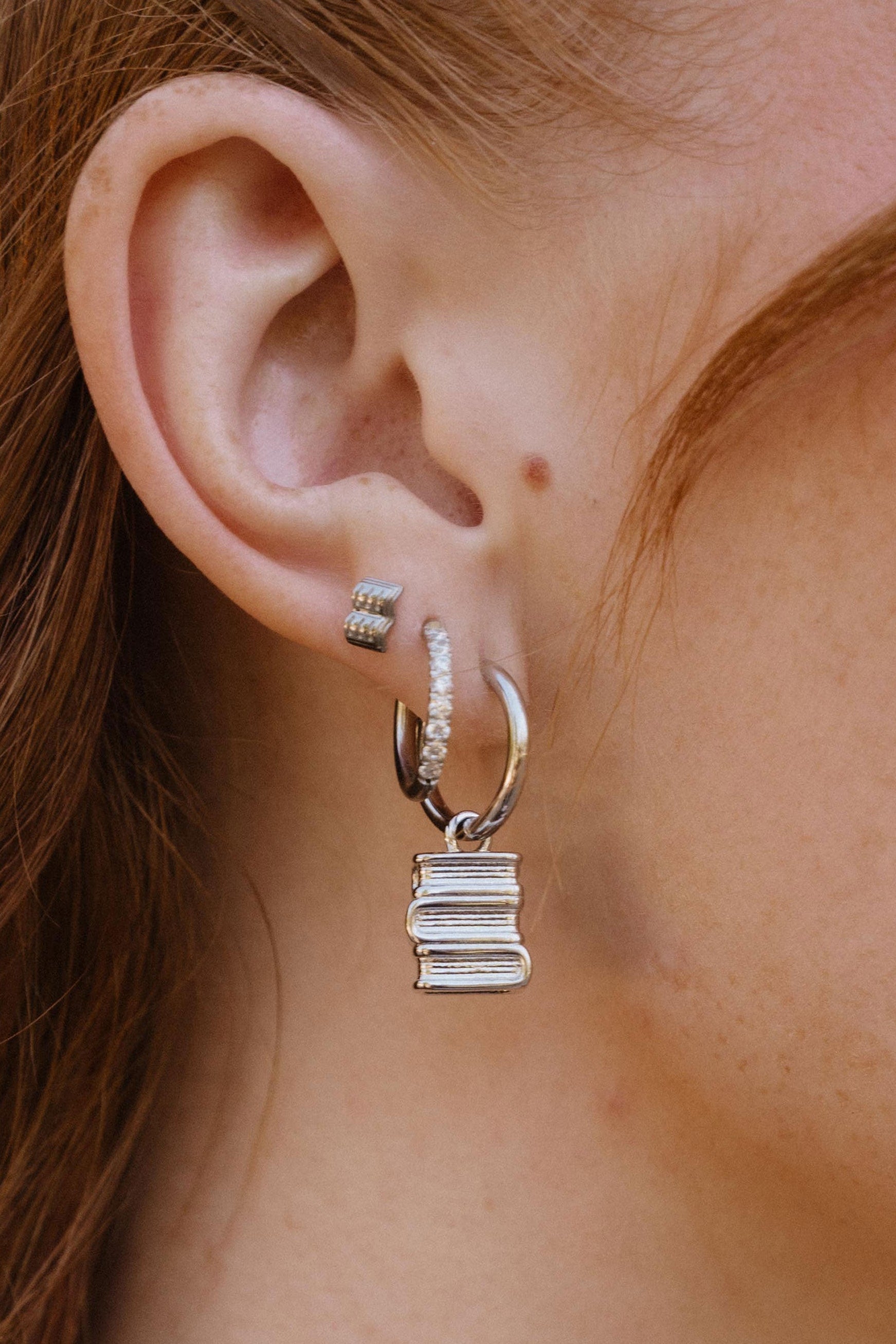 A close-up of a person's ear wearing multiple silver earrings, including a small stud, a huggie hoop with tiny clear stones, and a larger hoop with a dangling stacked books charm.