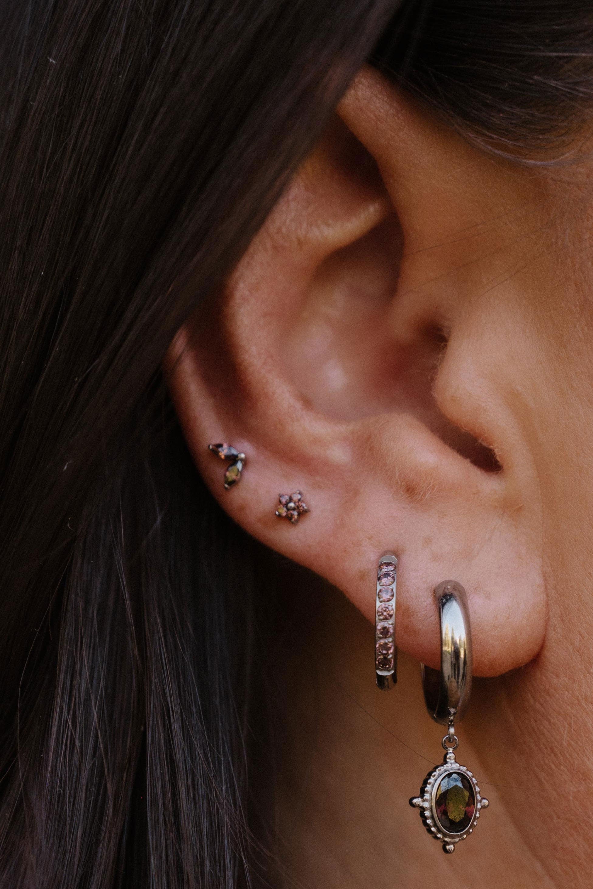Close-up of a person's ear showcasing multiple silver-toned piercings, including a hoop earring with a dangling oval charm, a hoop with multliple brown stones in a row, and two small floral studs.