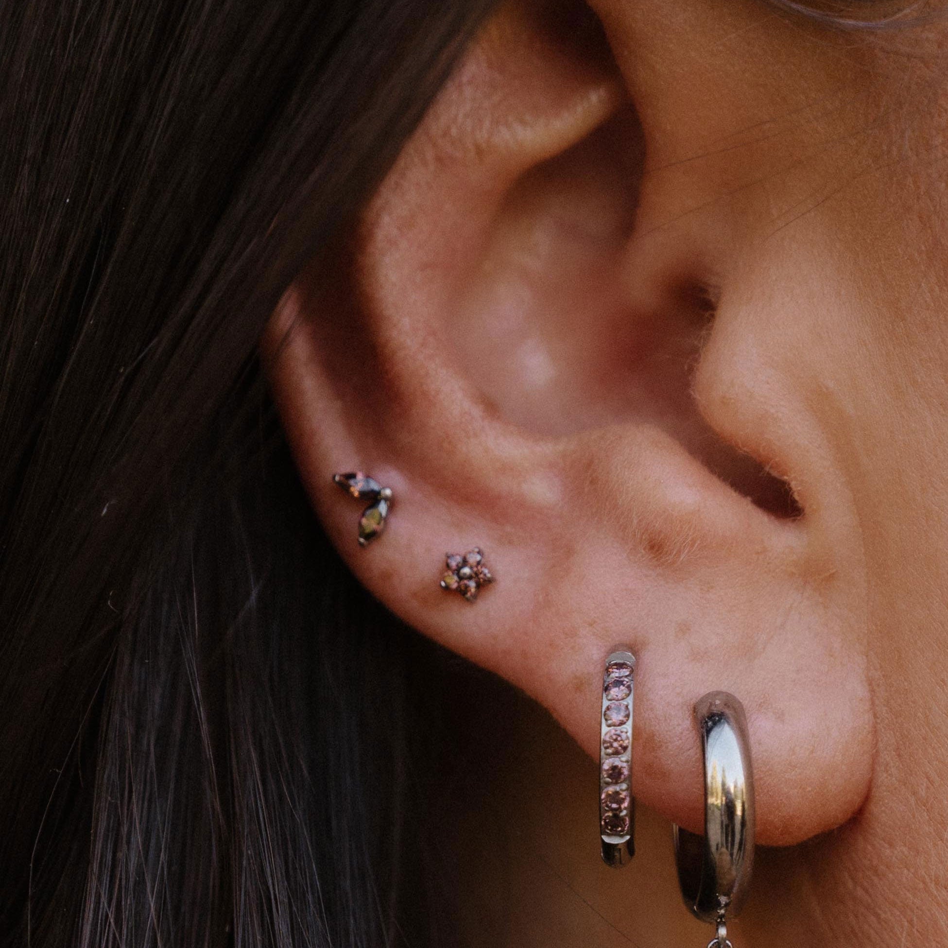 Close-up of a person's ear showcasing multiple silver-toned piercings, including a hoop earring with a dangling oval charm, a hoop with multliple brown stones in a row, and two small floral studs.