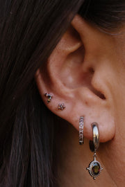 Close-up of a person's ear showcasing multiple silver-toned piercings, including a hoop earring with a dangling oval charm, a hoop with multliple brown stones in a row, and two small floral studs.