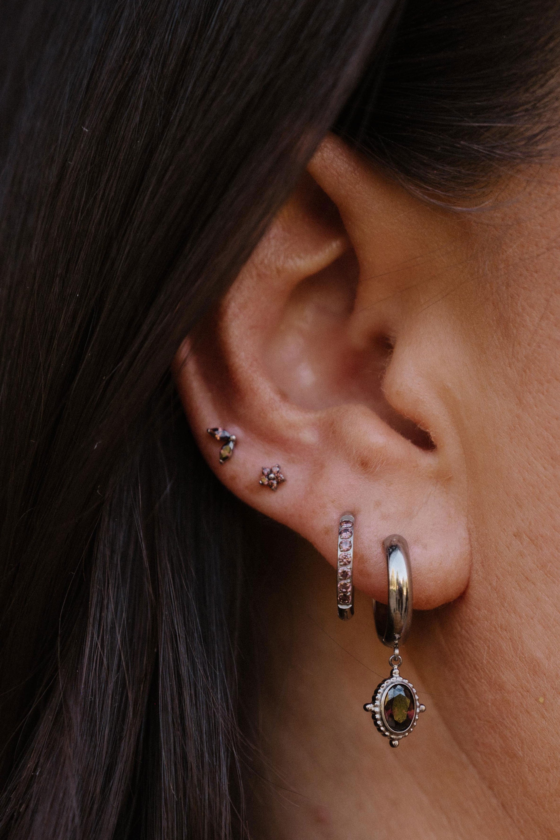 A close-up of a person's ear showcasing multiple silver-toned piercings, including a small hoop with brown stones, a dangling huggie with a round brown stone, and floral cartilage studs.