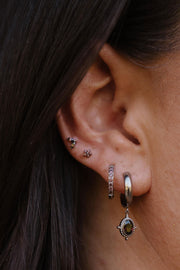 A close-up of a person's ear showcasing multiple silver-toned piercings, including a small hoop with brown stones, a dangling huggie with a round brown stone, and floral cartilage studs.