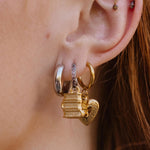 A close-up of a person's ear wearing a gold hoop earring with a small hanging gold charm that looks like a stack of three books, along with other ear piercings. The person has red hair.