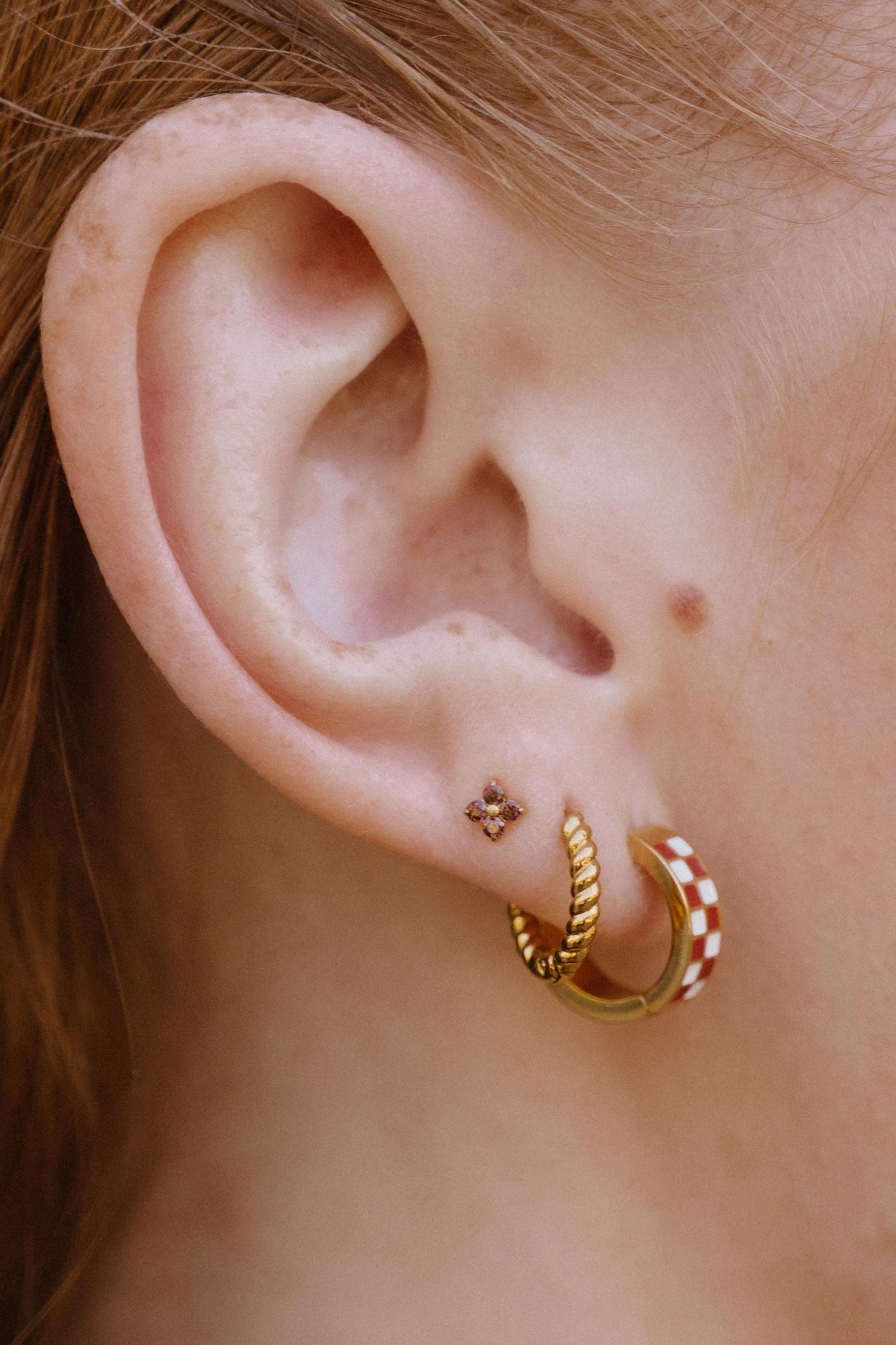 A close-up side profile of a person with reddish-brown hair wearing two small gold hoop earrings in their earlobe, one plain and one with brown and white checkered designs. In their third piercing is a brown four-gem stud.
