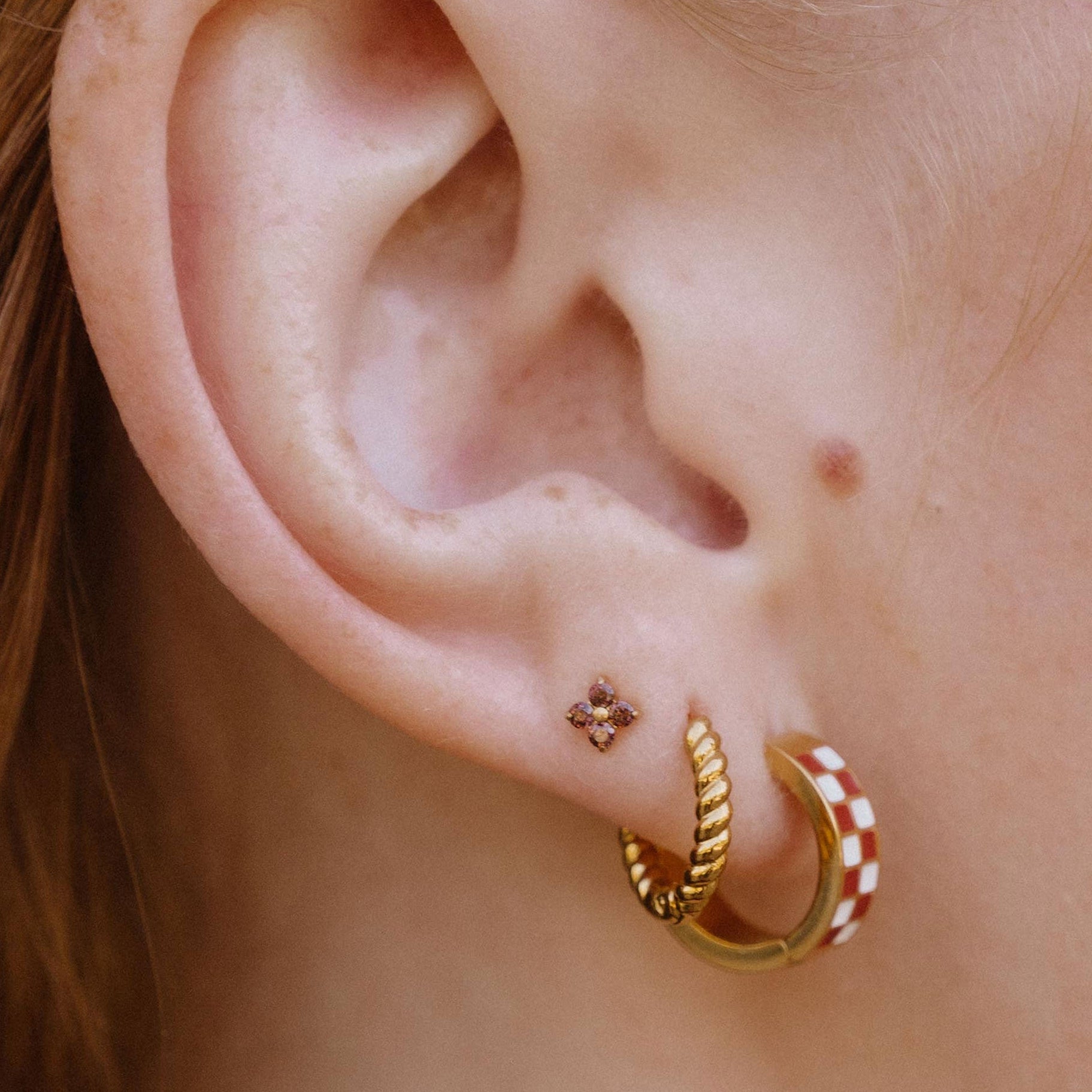 A close-up side profile of a person with reddish-brown hair wearing two small gold hoop earrings in their earlobe, one plain and one with brown and white checkered designs. In their third piercing is a brown four-gem stud.