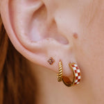 Close-up of a person's ear showcasing three gold hoop and stud earrings, including a red and white checkered hoop, a twisted gold rope hoop, and a small floral stud.