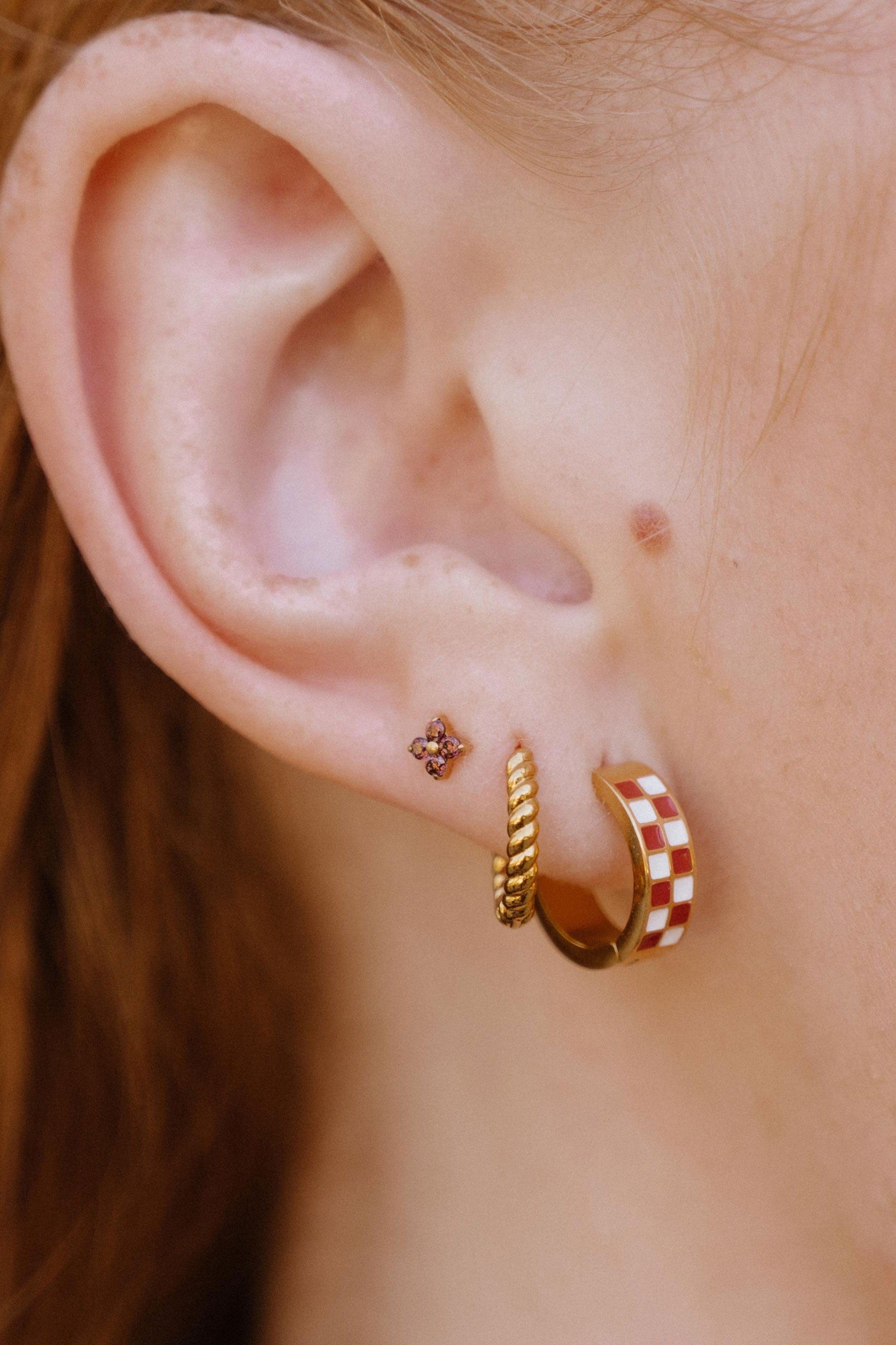Close-up of a person's ear showcasing three gold hoop and stud earrings, including a red and white checkered hoop, a twisted gold rope hoop, and a small floral stud.