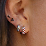 A close-up side profile of a person's ear wearing multiple small hoop and stud earrings, including a red-and-white checkered hoop and a silver hoop with a black dot stud.