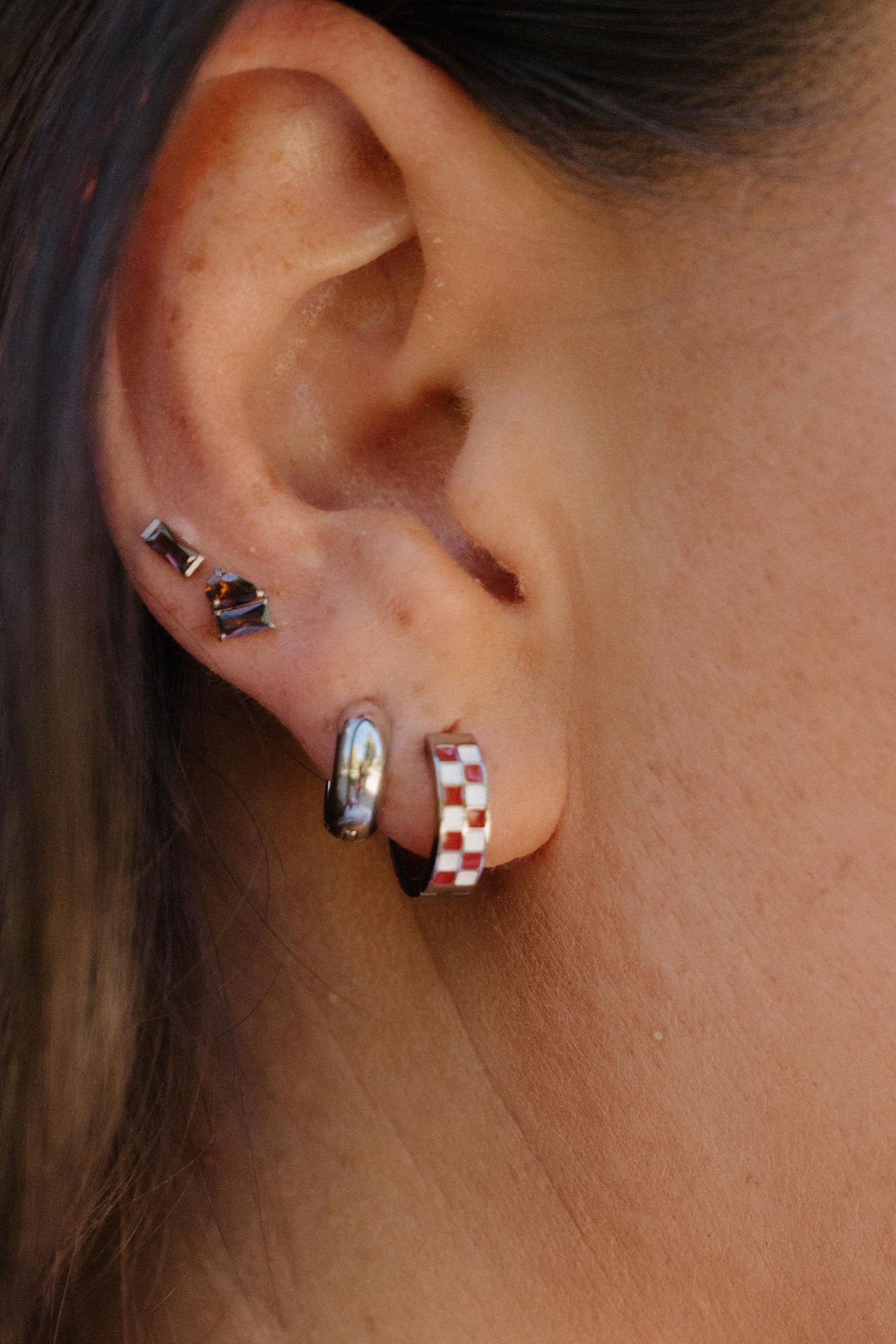 A close-up side profile of a person's ear wearing multiple small hoop and stud earrings, including a red-and-white checkered hoop and a silver hoop with a black dot stud.