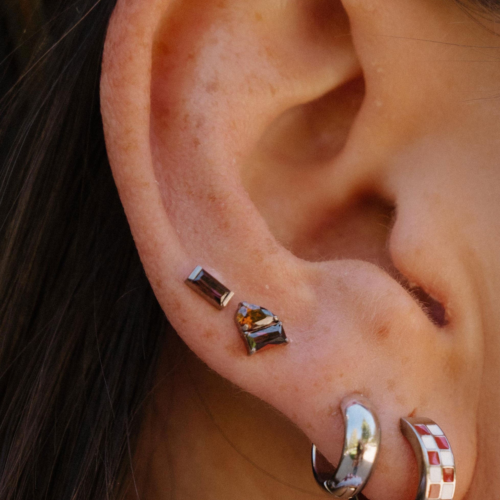 Close-up of an ear wearing multiple earrings, including two small stud earrings and two hoop earrings, one plain silver and one silver with a red and white checkered pattern.