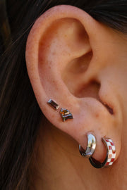 Close-up of an ear wearing multiple earrings, including two small stud earrings and two hoop earrings, one plain silver and one silver with a red and white checkered pattern.