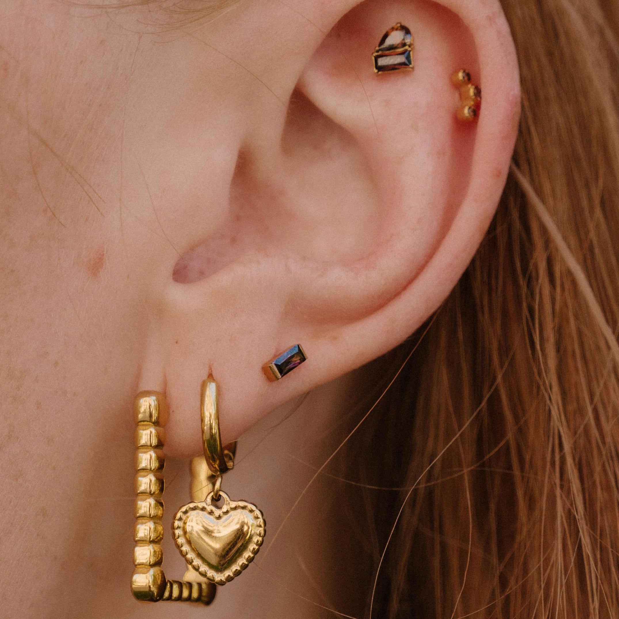 A close-up of a person's ear showcasing multiple gold piercings, including a large huggie hoop with a dangling beaded bar and heart charm, a small gold hoop, a rectangular dark blue stone stud, and ear cuffs.