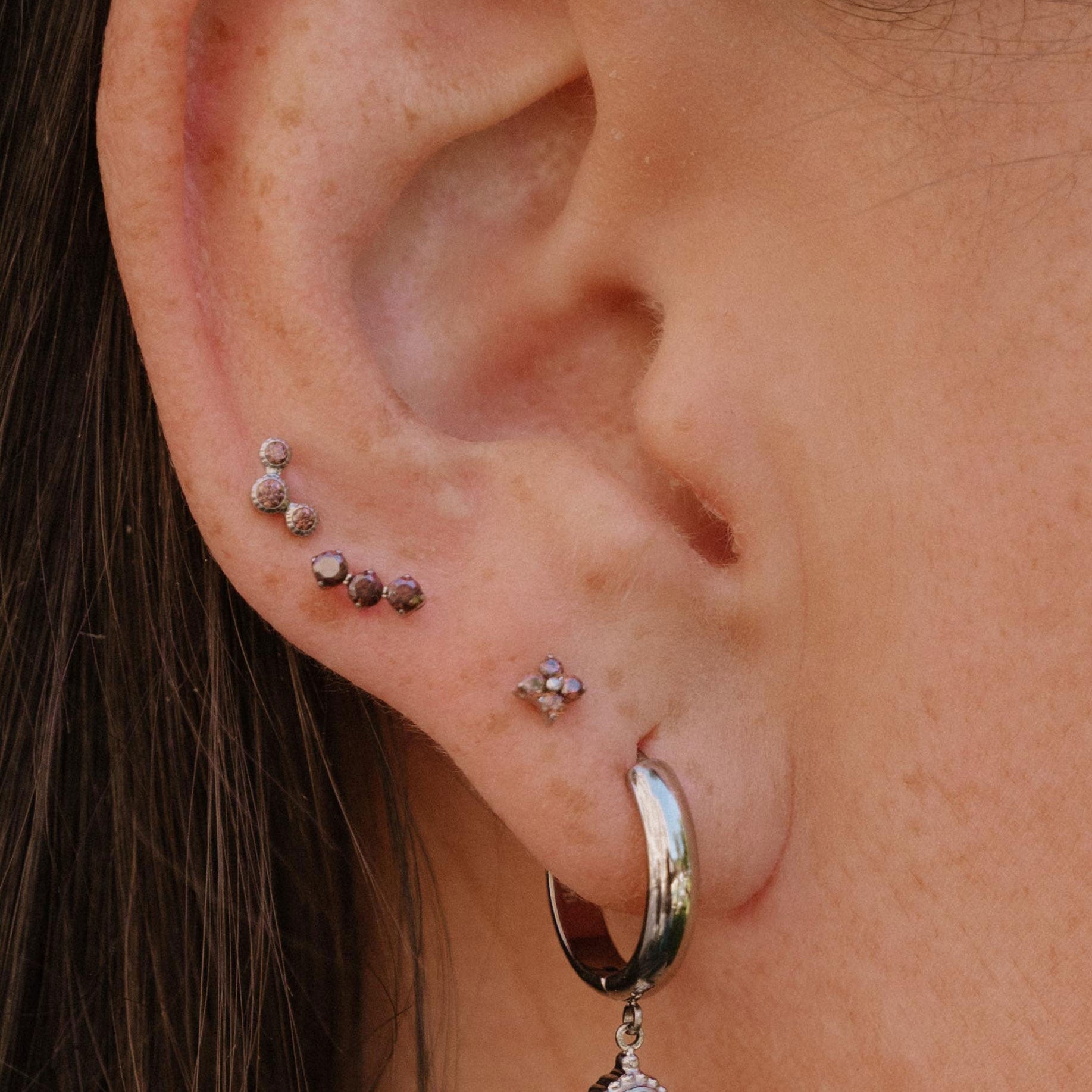 A close-up of a person's ear showcasing multiple piercings, including a silver hoop earring with a dangling brown stone and several small studs and piercings on the helix and lobe.