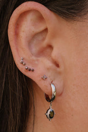 A close-up of a person's ear showcasing multiple piercings, including a silver hoop earring with a dangling brown stone and several small studs and piercings on the helix and lobe.
