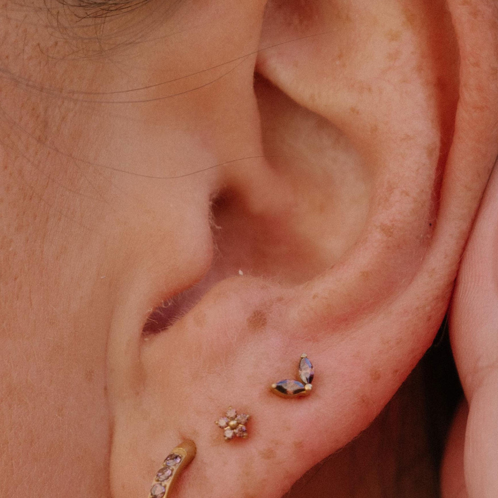 Close-up of a person's ear showcasing three gold-toned ear piercings: a curved hoop with small brown stones in the lobe, a small flower-shaped stud on the lobe, and a small two-leave brown stoned stud on the lobe.