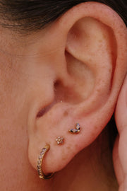 Close-up of a person's ear showcasing three gold-toned ear piercings: a curved hoop with small brown stones in the lobe, a small flower-shaped stud on the lobe, and a small two-leave brown stoned stud on the lobe.